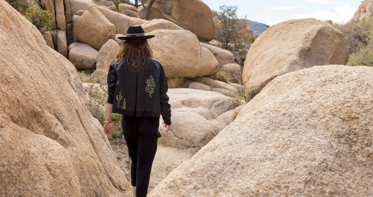 Dominique Cohen limited edition bomber jacket, black, with custom embroidery option, desert theme, on woman walking in Joshua Tree; call, text 323-404-2959 for help shopping our website or to make an appointment.